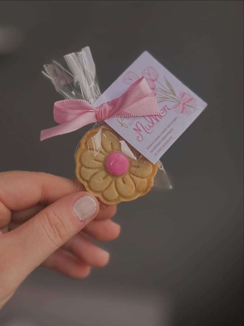 Biscoito decorado em formato de flor, embalado em saquinho de celofane com laço e tag personalizada, segurado na mão
